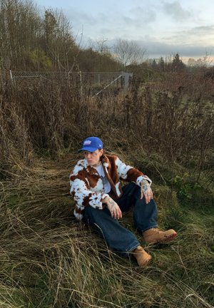 Persona con tatuajes que lleva una chaqueta con estampado de vaca, gorra azul, vaqueros y botas color tostado, sentada sobre hierba seca cerca de una valla de metal y árboles sin hojas.