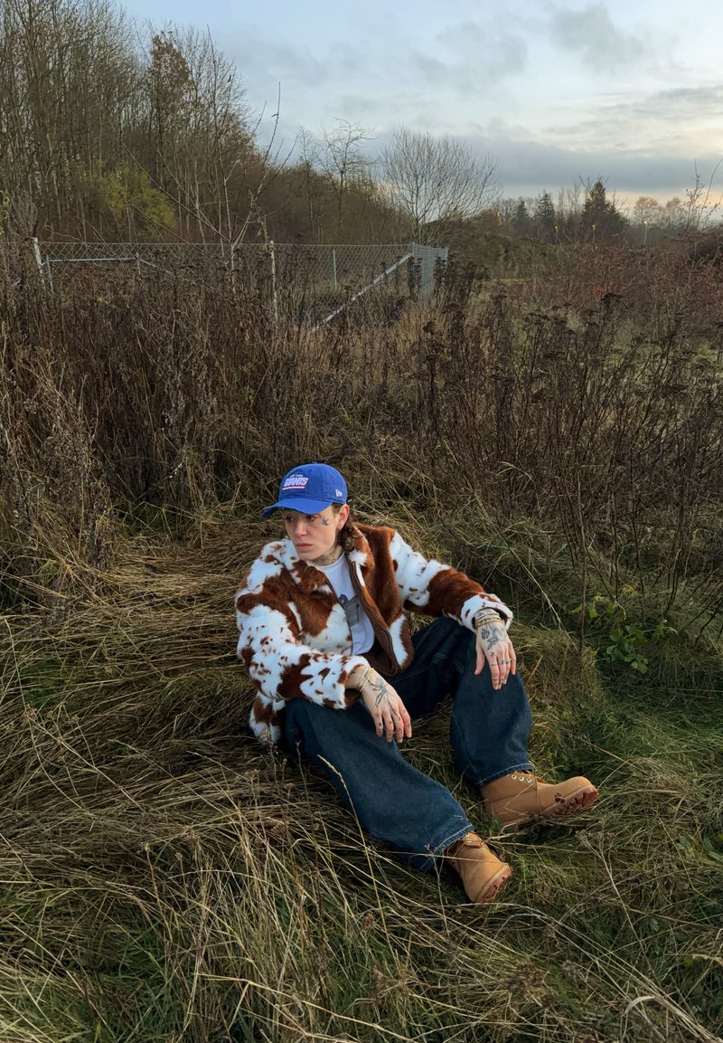Personne tatouée portant une veste à motif vache, une casquette bleue, un jean et des bottes beige, assise sur de l'herbe sèche près d'une clôture métallique et d'arbres sans feuilles.