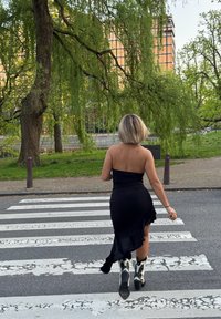 Czarna, bezramienna sukienka z opadającym dołem; widok z tyłu przedstawia modelkę w białych i czarnych, wzorzystych kowbojskich butach, przechodzącą przez ulicę w paski zebra.