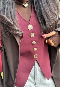 Femme portant un haut bordeaux boutonné avec cinq boutons dorés uniques, un sous-pull marron à manches longues, une veste marron et un pantalon gris, montrant une bague en or et des ongles roses.