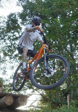 Bicicleta de montaña con un marco naranja, neumáticos negros y frenos de disco. El ciclista, que lleva una camiseta blanca y negra, realiza un salto sobre troncos de madera.