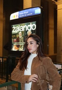 Brun mockajacka, vit topp och brun handväska med detaljrika metallbeslag, står nära en tunnelbaneskylt; nattmiljö med en reklam i bakgrunden.