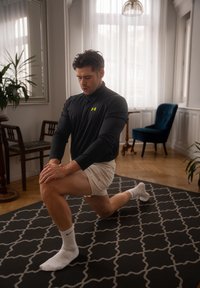 T-shirt de sport noir à manches longues avec logo néon, shorts blancs et chaussettes. Silhouette masculine en position de fente sur un tapis noir à motifs à l'intérieur.