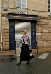 Person går på en gate, iført en rosa strikket topp med design av en svart katt, rutete ermer og et langt svart skjørt, holder en rosa håndveske.