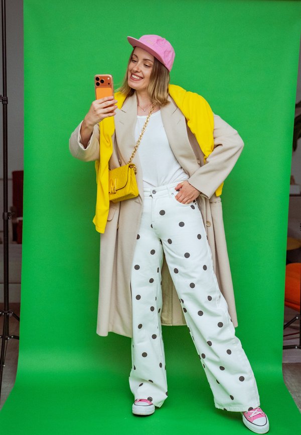 Donna con cappotto beige, maglione giallo, pantaloni bianchi a pois, cappellino rosa e scarpe rosa che scatta un selfie davanti a uno sfondo verde.