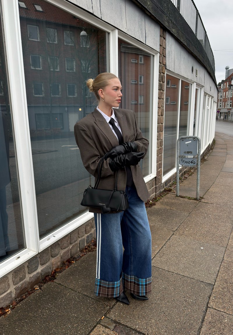 Femme en blazer oversize, chemise blanche, cravate noire, jeans larges avec revers à carreaux, gants noirs et sac à main, debout sur un trottoir en milieu urbain.