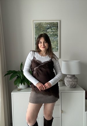 Joven con cabello castaño que lleva puesta una camisa blanca de manga larga, un camisón negro de encaje, una falda a cuadros y botas negras, de pie en el interior junto a un armario.