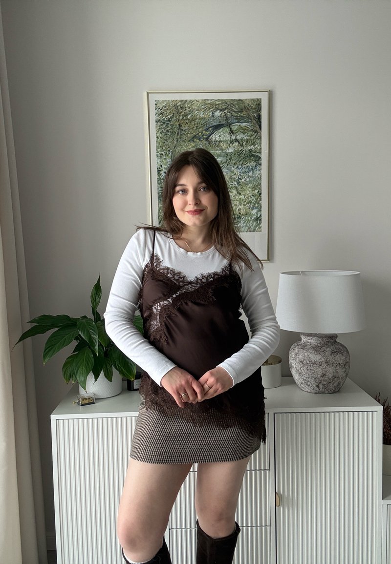 Jeune femme aux cheveux bruns portant une chemise blanche à manches longues, un caraco en dentelle noire, une jupe à carreaux et des bottes noires, debout à l'intérieur près d'un meuble.