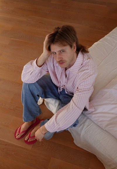Hombre sentado en el suelo cerca de la cama, vistiendo una camisa de rayas rosa, jeans azules y sandalias rojas, con una mano tocándose el cabello.