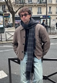 Νεαρός άνδρας με γυαλιά που φοράει μπεζ μπουφάν, καρό κασκόλ, ανοιχτόχρωμα τζιν και γάντια, στέκεται σε έναν δρόμο σε αστικό περιβάλλον.