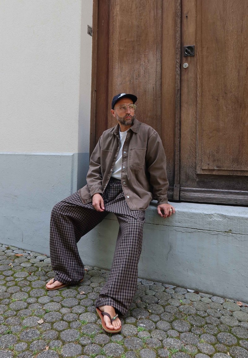 Homme portant une casquette, des lunettes, une veste marron, un pantalon large à carreaux et des tongs, assis sur un rebord en béton devant une porte en bois.