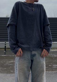 Personne portant une chemise bleu foncé à manches longues superposée, un jean bleu clair et un collier pendentif en forme de croix, debout à l'extérieur sur un trottoir mouillé.