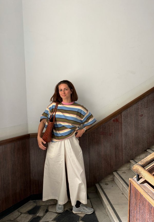 Femme debout sur un sol carrelé près des escaliers, portant une chemise rayée, un pantalon blanc ample, des baskets, et tenant un sac à bandoulière tressé marron.