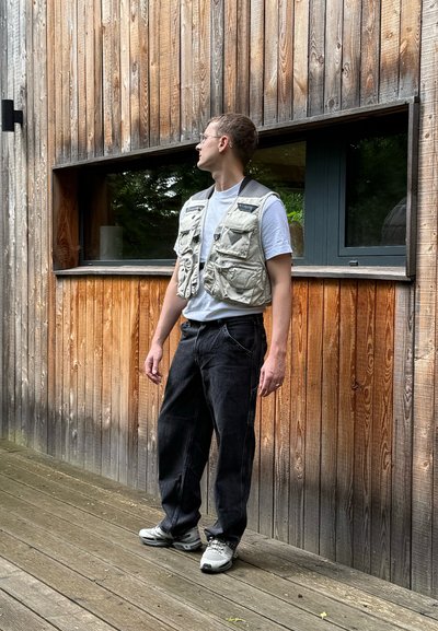 Chaleco de pesca color caqui con múltiples bolsillos, llevado sobre una camiseta blanca. Jeans de mezclilla oscura y zapatillas grises. Pared de madera en el fondo.