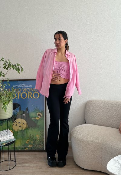 Camisa suelta de botón color rosa claro sobre un top corto fruncido rosa, combinada con pantalones de campana negros y zapatos negros gruesos. Planta verde en el fondo.