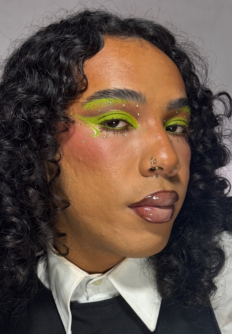 Person mit lockigem schwarzen Haar, die neon-grünen Lidschatten mit Strasssteinen trägt, glänzende dunkle Lippen, Nasenringe, weißes Hemd mit Kragen und schwarze Weste.