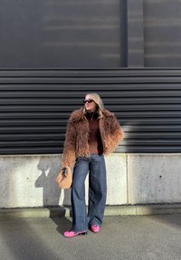 Femme portant une veste marron duveteuse, un jean large foncé, des chaussures roses à plateforme, des lunettes de soleil, tenant un sac à main texturé beige près d'un mur gris.
