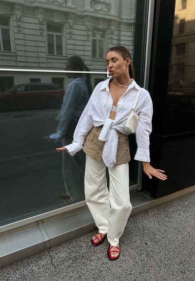 Camisa blanca de manga larga sobre un chaleco corto beige, combinada con pantalones blancos de pierna ancha y sandalias rojas de tiras, está de pie en una acera.