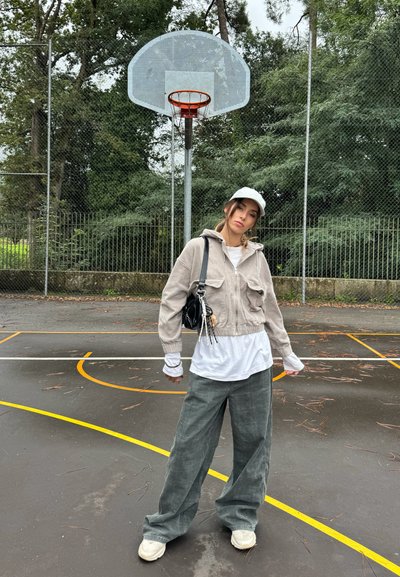Chaqueta de corte cropped de pana gris claro con bolsillos frontales, superpuesta a una camisa blanca. Pantalones de pierna ancha gris oscuro y zapatillas blancas. Fondo de cancha de baloncesto.