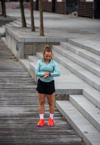 Sportowiec w jasnoniebieskiej koszulce z długim rękawem i czarnych szortach, sprawdzający zegarek fitness. Jasnopomarańczowe buty sportowe i białe skarpetki do kostek.