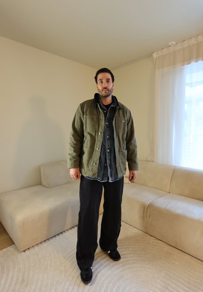 Hombre de pie en una sala de estar, vestido con una chaqueta verde oliva, camisa oscura, pantalones negros holgados y zapatos negros; sofá beige y cortina clara al fondo.