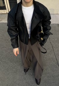 Veste courte en cuir noir avec larges revers, chemise blanche ajustée en dessous, associée à un pantalon ample marron et des chaussures noires.