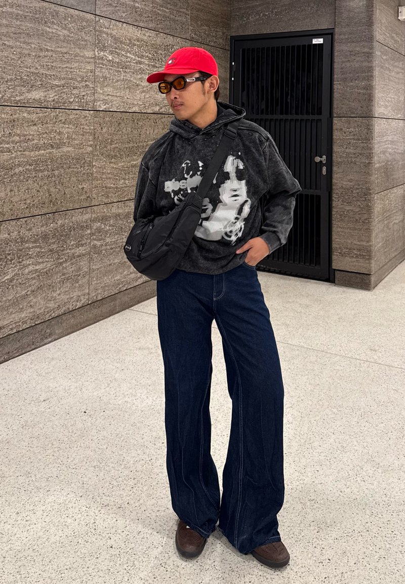 Sweat à capuche gris avec impression blanche, large jean en denim bleu, sac bandoulière noir, casquette rouge et chaussures marron avec semelles texturées.