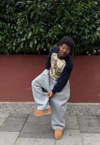 Pantalon de survêtement gris surdimensionné, pull noir côtelé avec graphique, bottes beiges, debout sur un pavé en pierre devant une haie verte et un mur rouge.