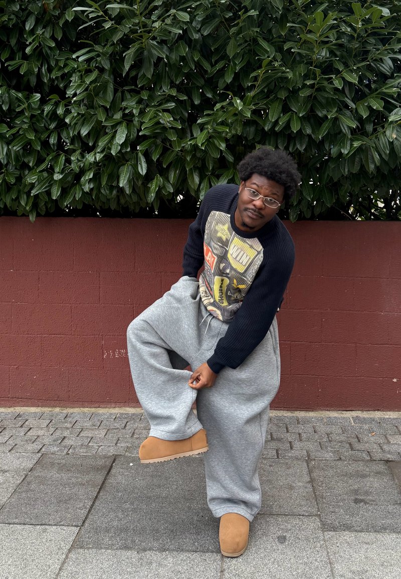 Pantaloni sportivi grigi oversize, maglione nero a coste con grafica, stivali beige, in piedi su pavimento in pietra davanti a una siepe verde e a un muro rosso.