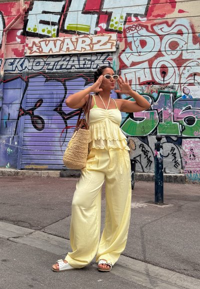 Top peplum arrugado de color amarillo claro combinado con pantalones amarillos de pierna ancha, bolso de mimbre y sandalias blancas. Fondo de graffiti colorido.