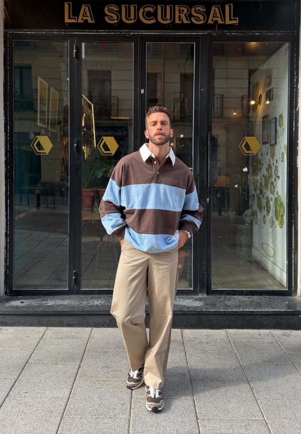 Homme portant une chemise rayée marron et bleu clair avec un pantalon beige et des baskets, debout les mains dans les poches devant une vitrine noire avec le panneau "La Sucursal".