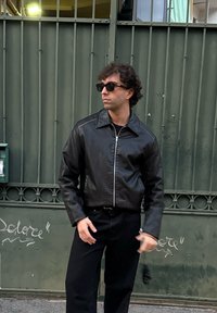 Μαύρη δερμάτινη ζακέτα με φερμουάρ, φαρδύ γιακά και μακριά μανίκια, σε συνδυασμό με μαύρο παντελόνι, μπροστά σε πράσινη πύλη.