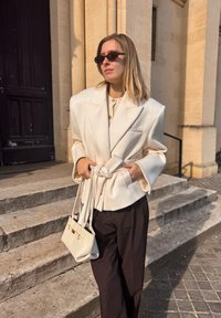 Krémové štruktúrované sako so širokými klopami, zviazané v páse, v kombinácii s tmavými voľnými nohavicami a bielou kabelkou so zlatými doplnkami.