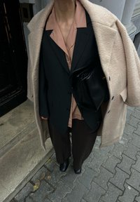 Personne portant un manteau beige par-dessus un blazer noir, un pantalon ample marron, des bottes noires, et tenant un sac en cuir noir sur un trottoir pavé.