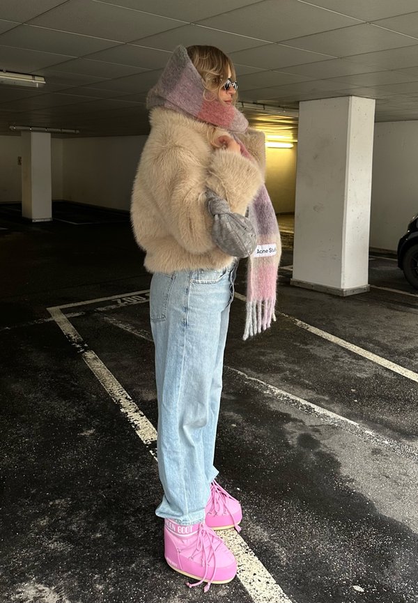 Veste en fausse fourrure beige, écharpe pastel superposée, jeans droit bleu clair, et bottes à lacets roses avec accents de logo.
