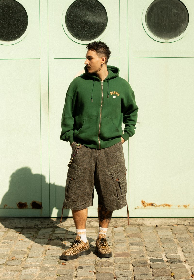 Mężczyzna stojący na kostkowej ulicy w zielonym rozpinanym hoodie, wzorzystych szortach cargo i butach w leopardzie wzory, spoglądający w bok na jasnzielone drzwi.