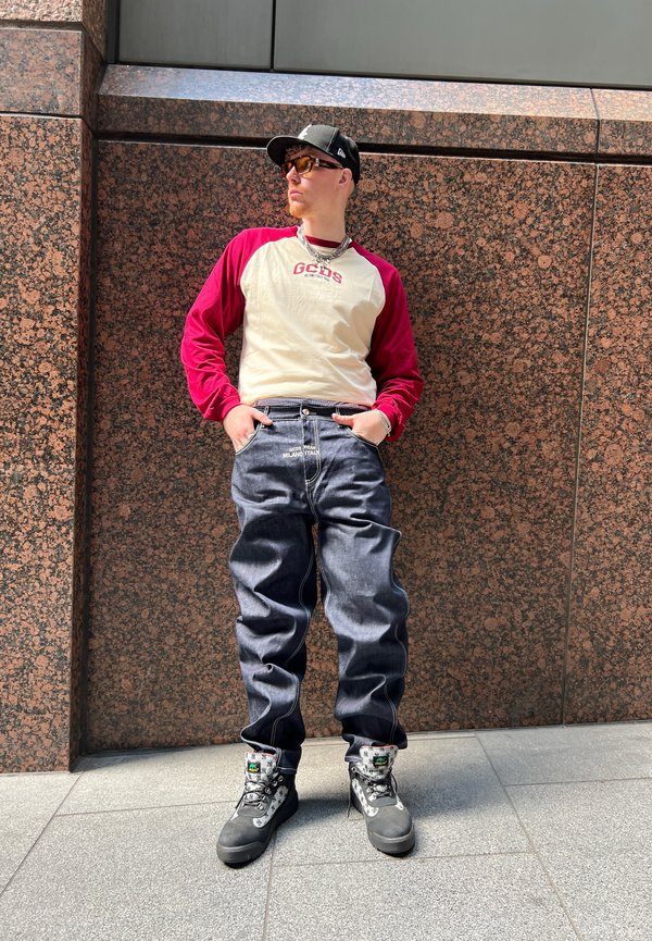 Joven vestido con una camiseta de manga larga blanca y roja, jeans holgados, botas de senderismo negras, gafas de sol y una gorra negra, de pie contra una pared de mármol marrón.