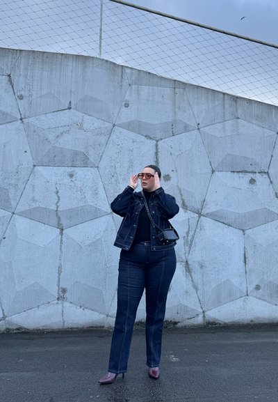 Chaqueta de mezclilla y jeans a juego, ambos de un azul oscuro, combinados con una blusa negra ajustada. Gafas de sol claras y tacones burdeos brillantes completan el look.