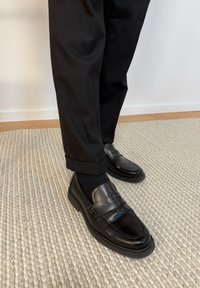 Μαύρα δερμάτινα μοκασίνια φορεμένα με μαύρες κάλτσες και μαύρο παντελόνι με μανσέτες, που στέκονται πάνω σε μπεζ υφασμάτινο χαλί.