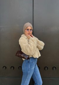 Krämfärgad fuskpälsjacka, ljusbrun stickad mössa, blå jeans med hög midja och mörkbrun axelväska mot en grå bakgrund.