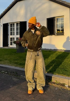 Chaqueta bomber de pana color oliva con un logo, jeans oversized de color verde claro, gorro amarillo y calzado marrón. De pie frente a una casa blanca.