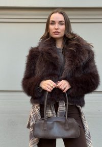 Jeune femme portant un manteau en fourrure marron foncé et des bagues, tenant un sac à main en cuir marron foncé contre un mur beige.