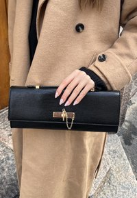 Čierna textúrovaná clutch kabelka so zlatým reťazovým akcentom a zapínaním, obsahujúca priehľadný prvok. Drží ju ruka, blízko béžového kabáta.