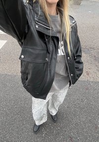 Veste de moto en cuir noir avec fermetures éclair argentées et détails de poche, associée à un sweat-shirt gris et un pantalon large à motifs.