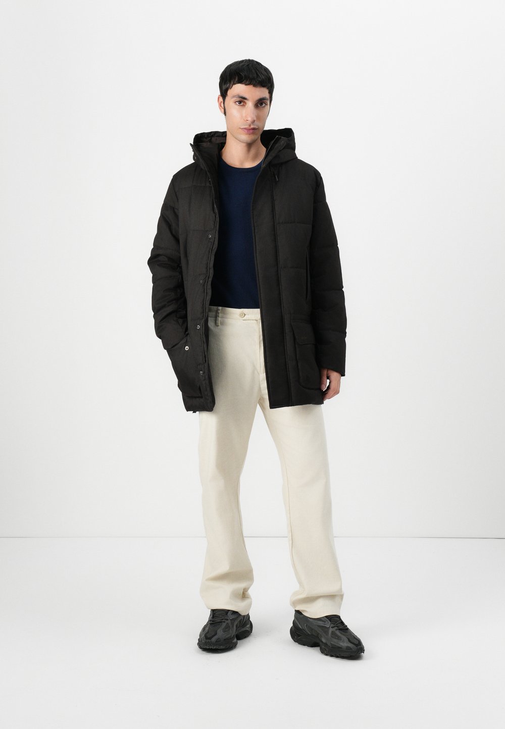 Fekete pufferszövet kabát kapucnival, tűzött mintával �és két elülső zsebbel. Tengerészkék ing fölött viselve, krémszínű nadrággal és fekete cipővel párosítva.