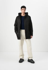 Fekete pufferszövet kabát kapucnival, tűzött mintával és két elülső zsebbel. Tengerészkék ing fölött viselve, krémszínű nadrággal és fekete cipővel párosítva.