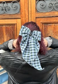 Fascia a fiocco a quadri blu e verdi con un centro nero, fissata a capelli scuri, su uno sfondo di legno. Giacca di pelle visibile.