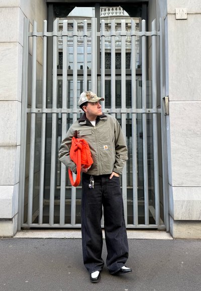 Hombre con gorra de camuflaje, chaqueta gris y pantalones anchos negros sostiene una bolsa naranja brillante frente a una puerta metálica con rejas.