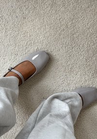 Błyszczące szare buty Mary Jane o kwadratowych noskach i pasku na kostce, w połączeniu z jasnoszarymi, luźnymi spodniami na teksturowanym dywanie.