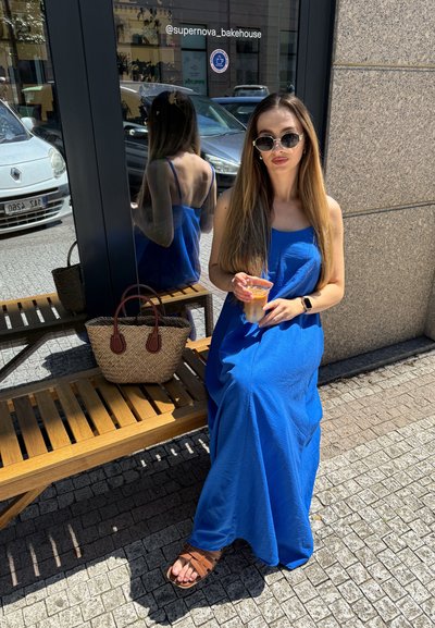 Vestido maxi de color azul royal, bolsa tejida con asas marrones, vaso transparente con una bebida, sentado en un banco de madera contra una pared de piedra.